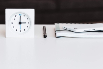 Image shows an analogue clock on a white desk beside a pen, notebook and calculator.