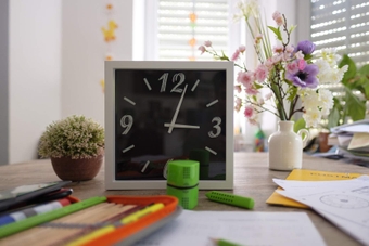 Image shows a desk with an analogue clock, stationary, and flowers.